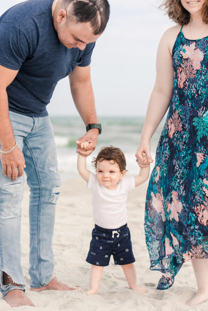 couple holding infant son's hands while trying to walk on the beach