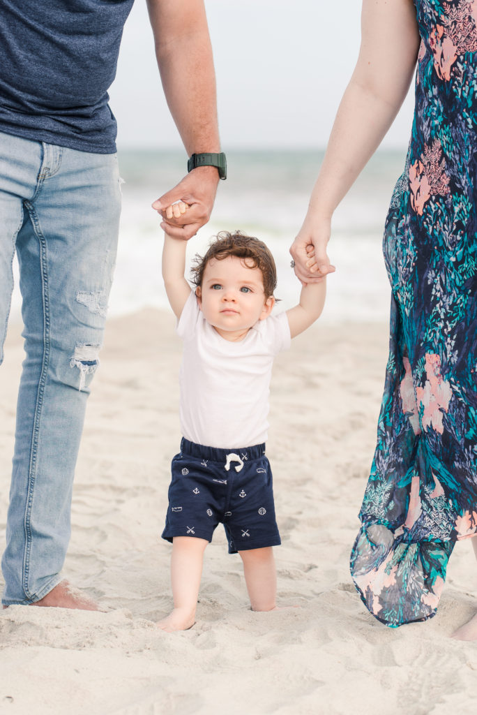 infant holding parents hands while trying to stand during family beach session
