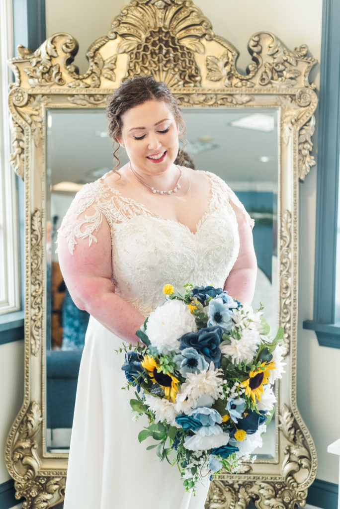 bride holding blue yellow and white wedding bouquet in front of large ornate mirror