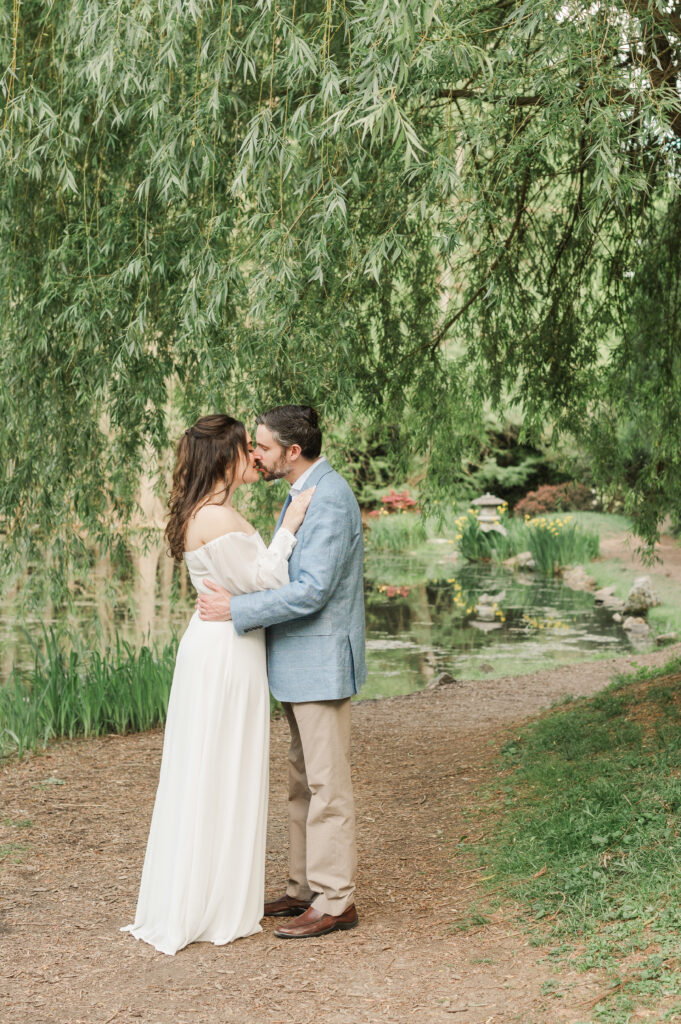 engaged couple kissing in front of willow tree on gravel path