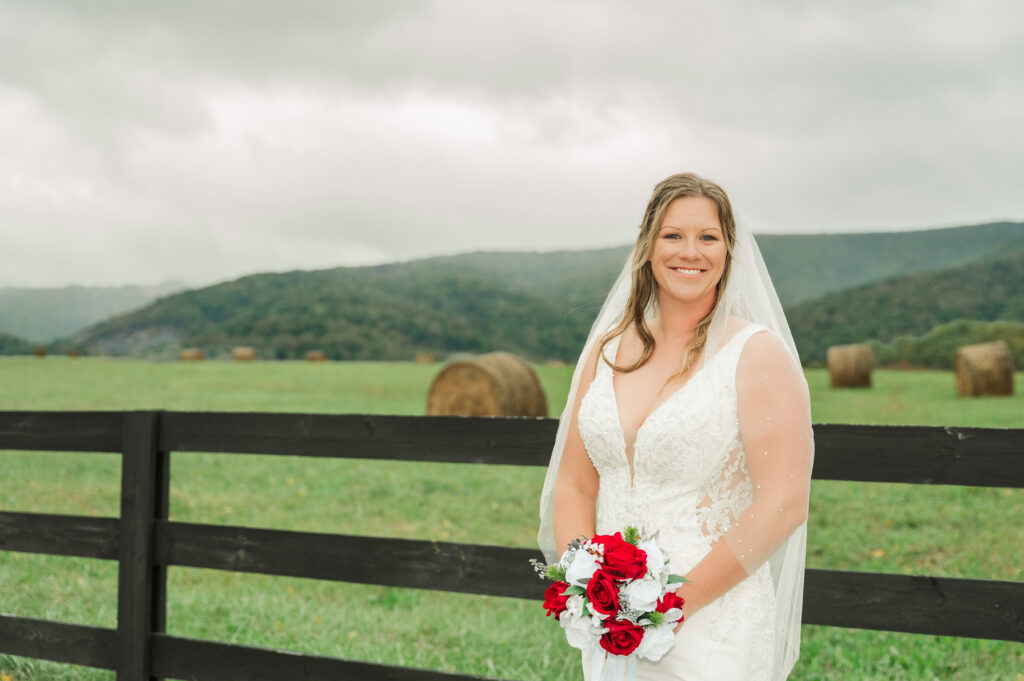 A bride in a flowing white wedding gown standing by a black farm fence with the Blue Ridge Mountains and round hay bales in the background.