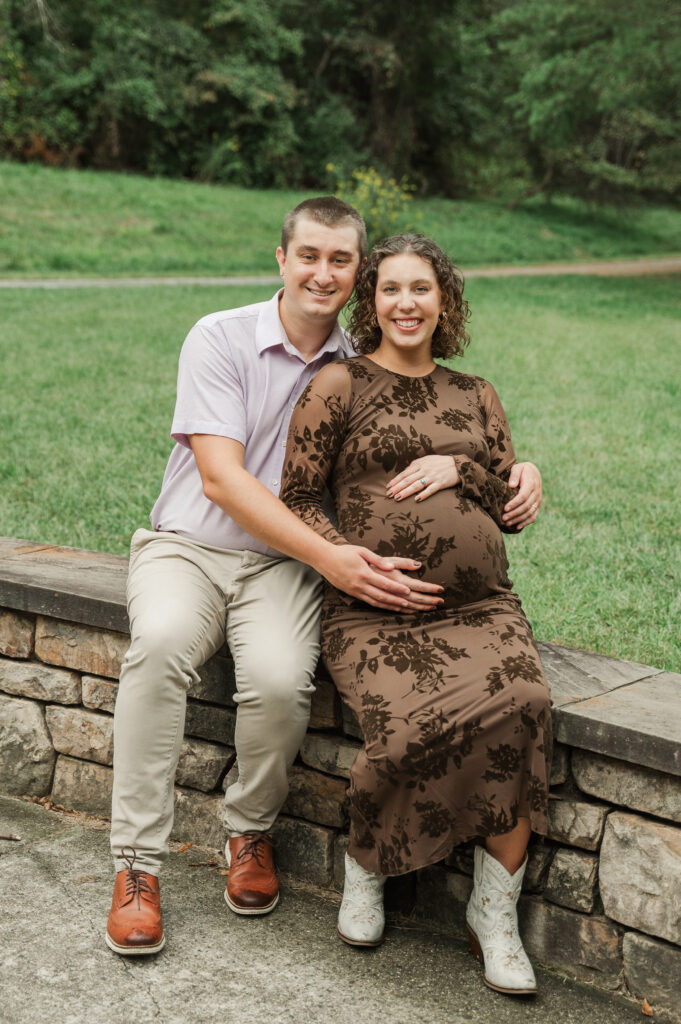 Pregnant Emily and husband Derek sitting together on a long rock wall on the Saunders-Monticello Trail, smiling at the camera