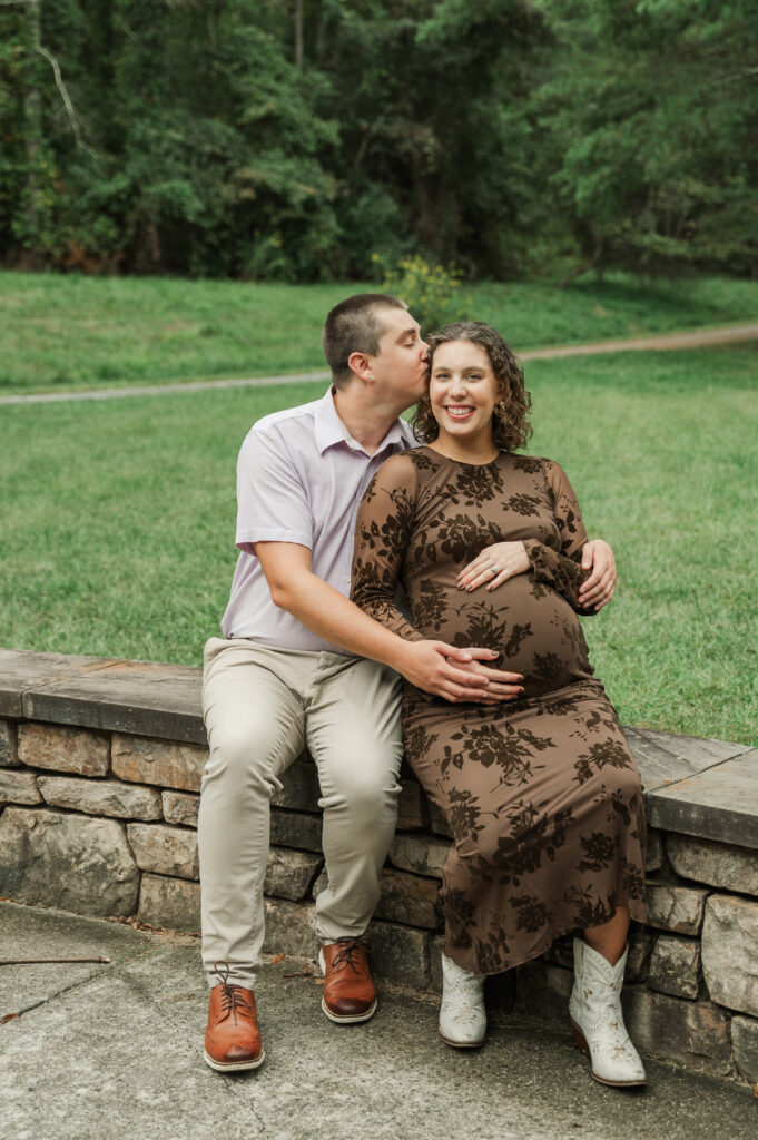 Full-length photo of Emily and Derek sitting comfortably on a rock wall, showing off her fitted dress and white boots