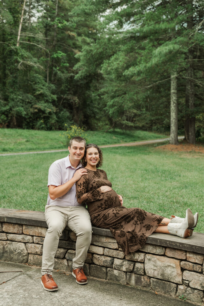Candid maternity portrait of Emily sitting on a rock wall, with her resting her hand on her baby bump against a lush, cloudy trail backdrop