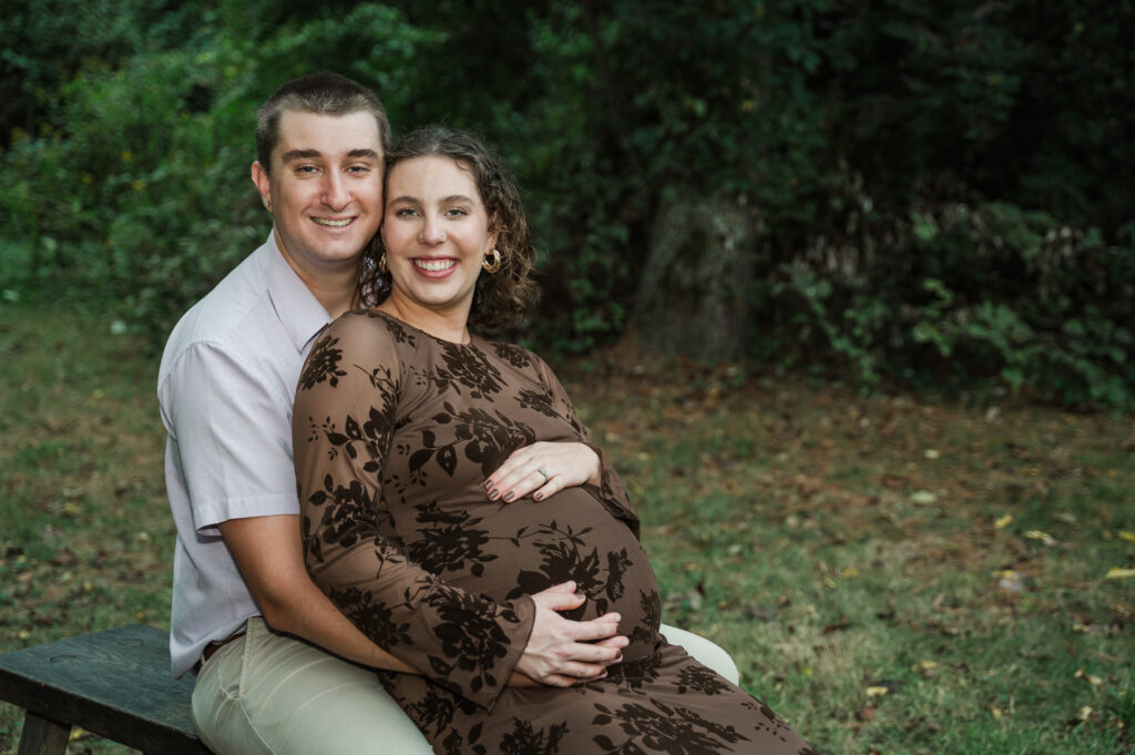 Candid maternity portrait of Emily sitting on a bench with Derek resting his hand on her baby bump ]